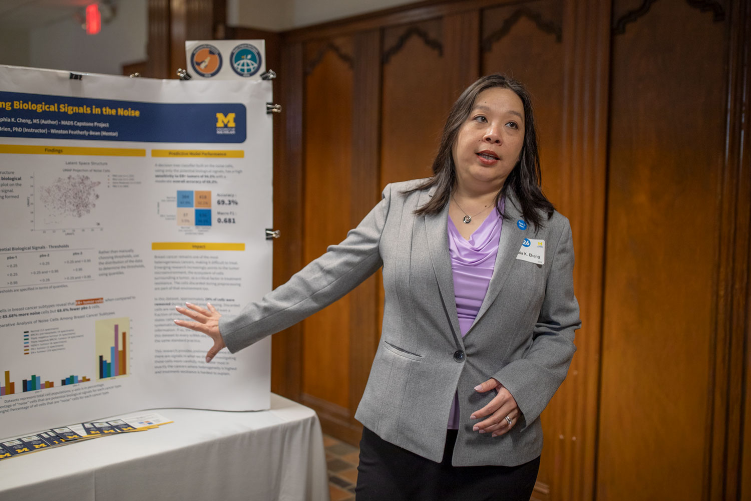 Woman in a gray blazer presents a research poster at an academic conference, gesturing toward charts and data on a display board.