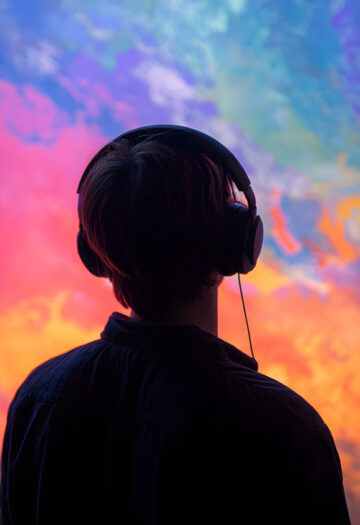 Person wearing over-ear headphones viewed from behind, facing a vivid abstract cloudscape of swirling pink, blue, purple, and orange colors.