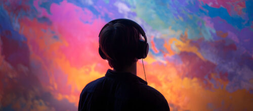 Person wearing over-ear headphones viewed from behind, facing a vivid abstract cloudscape of swirling pink, blue, purple, and orange colors.