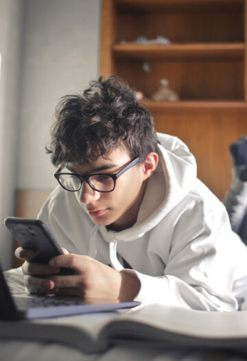 A young person wearing glasses and a white hoodie lies on a bed, looking at a smartphone beside an open laptop.