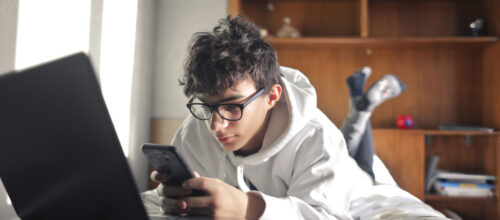 A young person wearing glasses and a white hoodie lies on a bed, looking at a smartphone beside an open laptop.