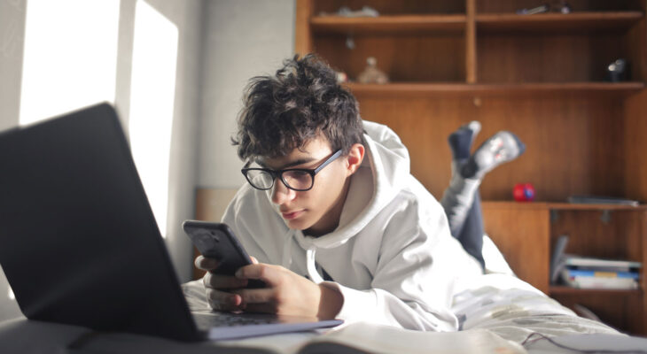 A young person wearing glasses and a white hoodie lies on a bed, looking at a smartphone beside an open laptop.