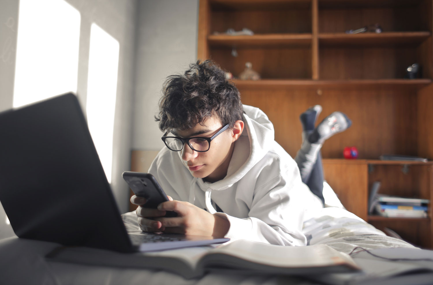 A young person wearing glasses and a white hoodie lies on a bed, looking at a smartphone beside an open laptop.