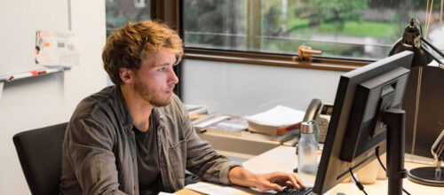 A man sits at a desk, focused on a desktop computer in a bright office with large windows behind him.