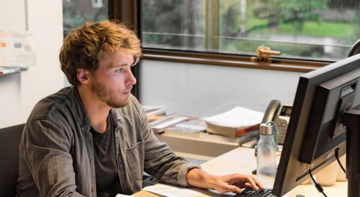 A man sits at a desk, focused on a desktop computer in a bright office with large windows behind him.