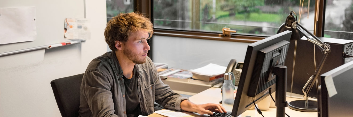 A man sits at a desk, focused on a desktop computer in a bright office with large windows behind him.