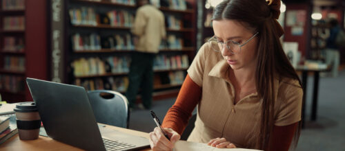 Woman sitting at desk in library writing in a notebook with a laptop next to her.