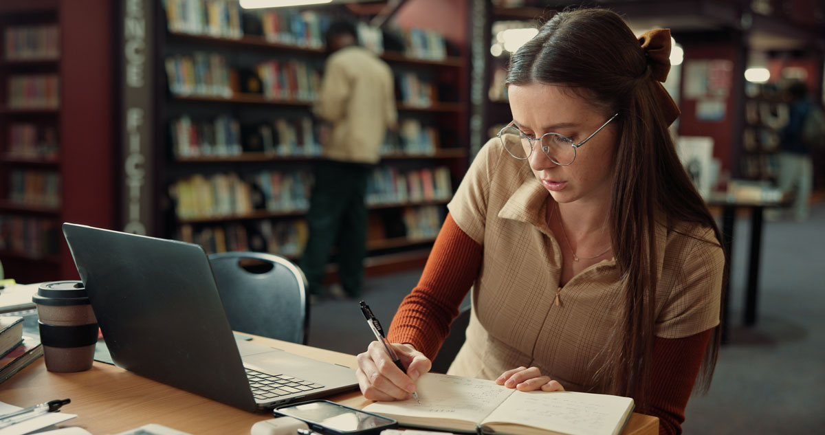 Woman sitting at desk in library writing in a notebook with a laptop next to her.