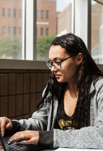 A person with long dark hair and glasses types on a sticker-covered laptop while seated at a table in a bright building with large windows.