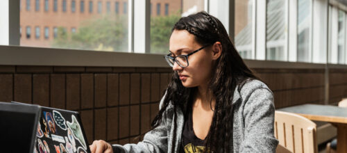 A person with long dark hair and glasses types on a sticker-covered laptop while seated at a table in a bright building with large windows.