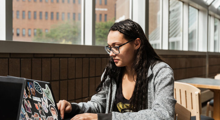 A person with long dark hair and glasses types on a sticker-covered laptop while seated at a table in a bright building with large windows.