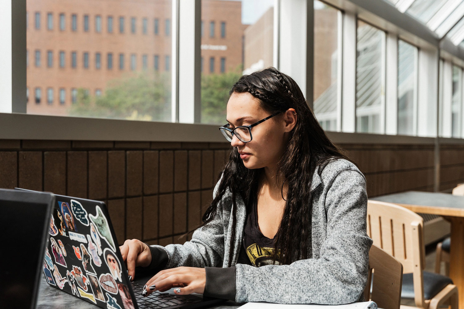 A person with long dark hair and glasses types on a sticker-covered laptop while seated at a table in a bright building with large windows.