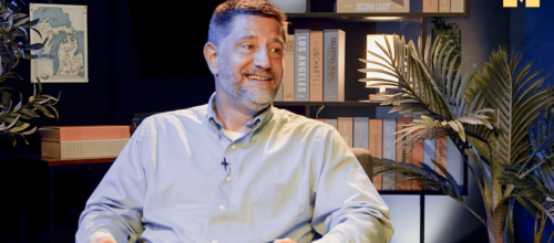 A smiling man in a light blue button-down shirt sits in an armchair in a warmly lit studio.