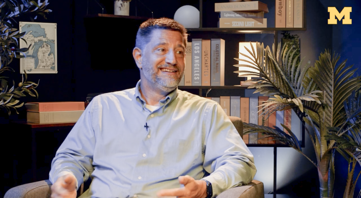 A smiling man in a light blue button-down shirt sits in an armchair in a warmly lit studio.