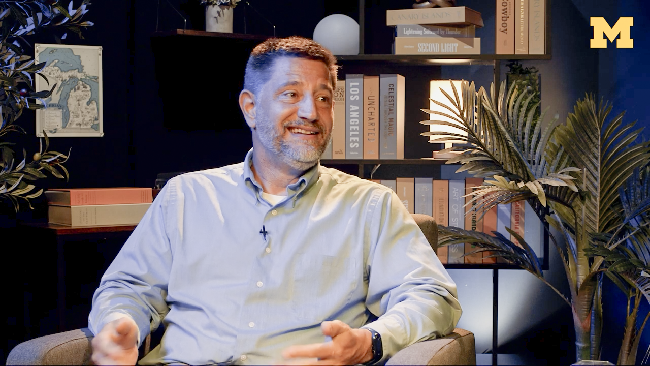 A smiling man in a light blue button-down shirt sits in an armchair in a warmly lit studio.