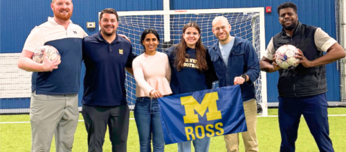 Six people pose together on an indoor soccer field in front of a goal, with two holding soccer balls and two holding a blue-and-maize Michigan Ross flag.