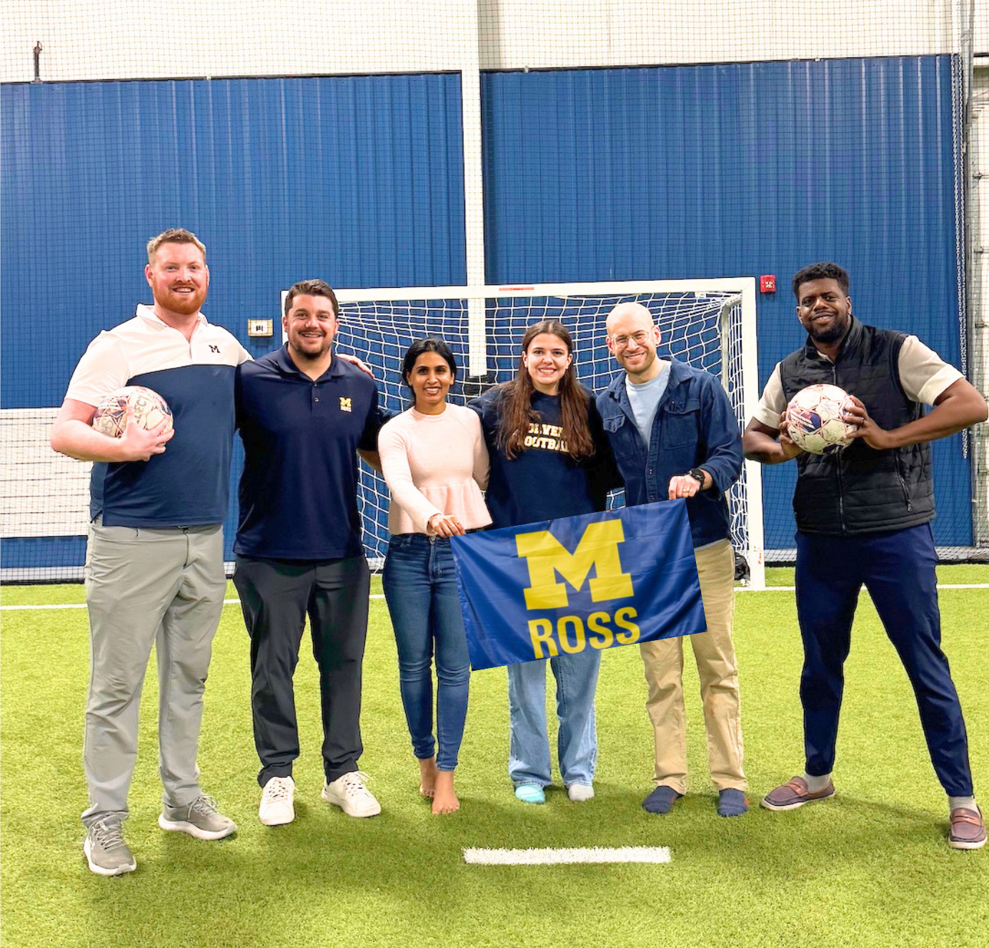 Six people pose together on an indoor soccer field in front of a goal, with two holding soccer balls and two holding a blue-and-maize Michigan Ross flag.
