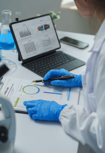 A scientist in a white lab coat and blue protective gloves examines printed research charts while holding a pen. A tablet displays multiple graphs, with a microscope, laboratory glassware, and containers of blue liquid arranged across the white workbench.