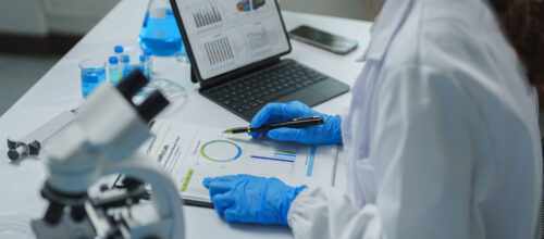 A scientist in a white lab coat and blue protective gloves examines printed research charts while holding a pen. A tablet displays multiple graphs, with a microscope, laboratory glassware, and containers of blue liquid arranged across the white workbench.