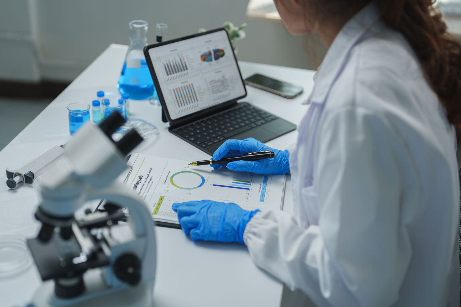A scientist in a white lab coat and blue protective gloves examines printed research charts while holding a pen. A tablet displays multiple graphs, with a microscope, laboratory glassware, and containers of blue liquid arranged across the white workbench.