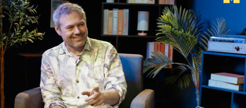 A smiling gray-haired man wearing a light patterned shirt sits in an armchair and gestures while looking to his right. Bookshelves, leafy plants, and blue accent lighting create a relaxed studio setting, with a yellow University of Michigan block “M” in the upper-right corner.