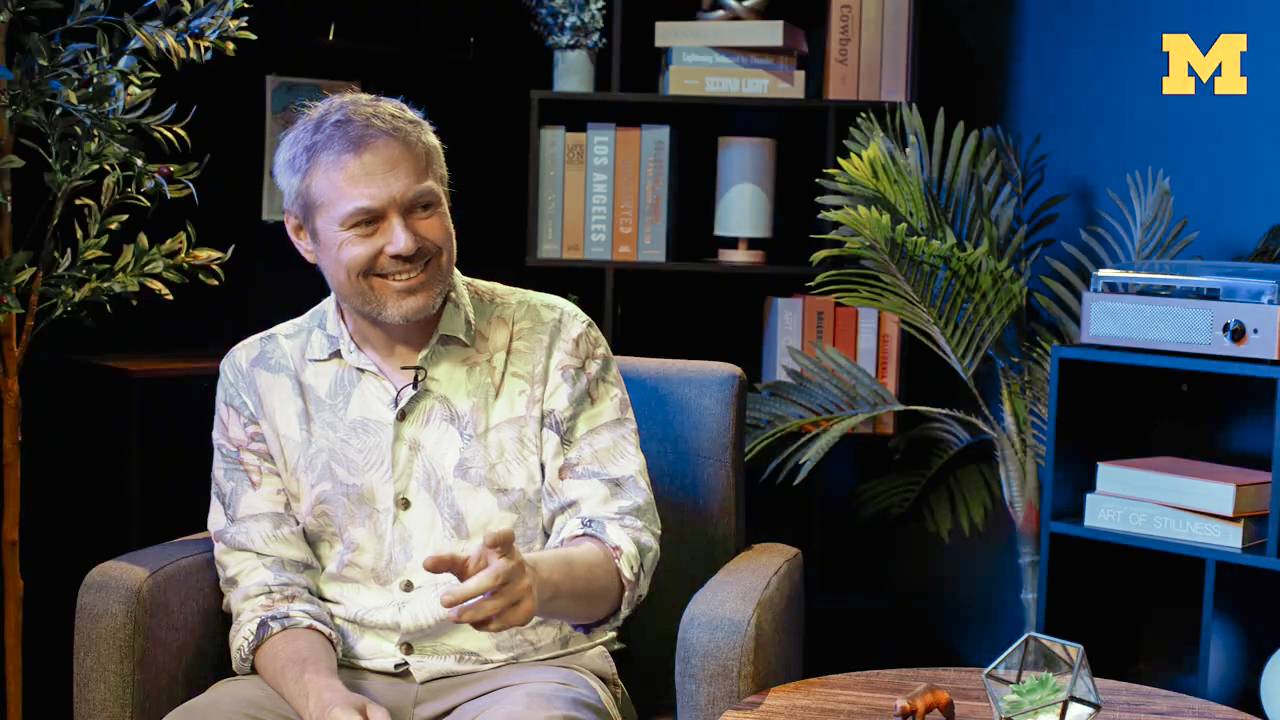 A smiling gray-haired man wearing a light patterned shirt sits in an armchair and gestures while looking to his right. Bookshelves, leafy plants, and blue accent lighting create a relaxed studio setting, with a yellow University of Michigan block “M” in the upper-right corner.